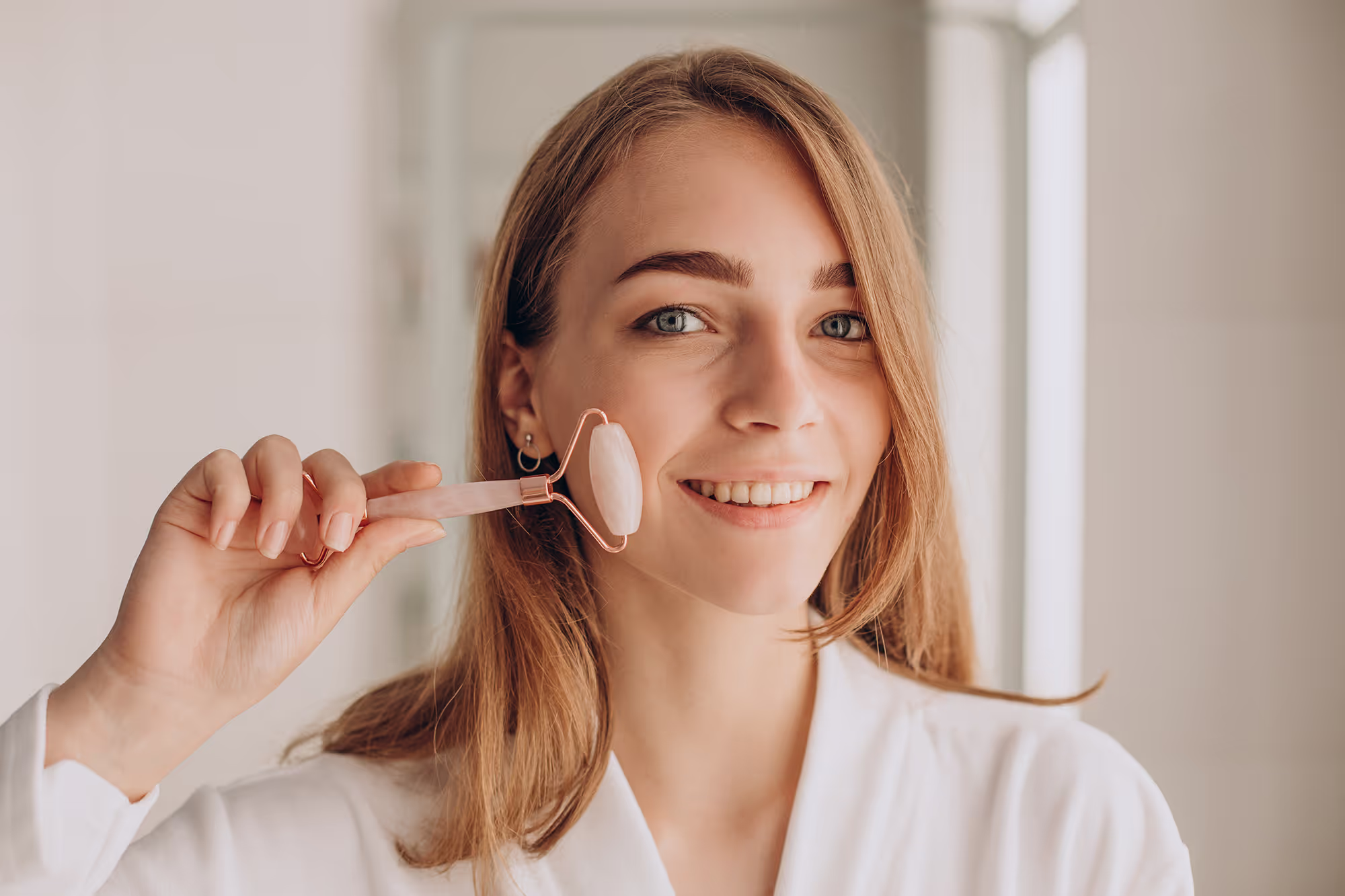 Smiling woman holding a rose quartz face roller, using it for a self-massage on her face
